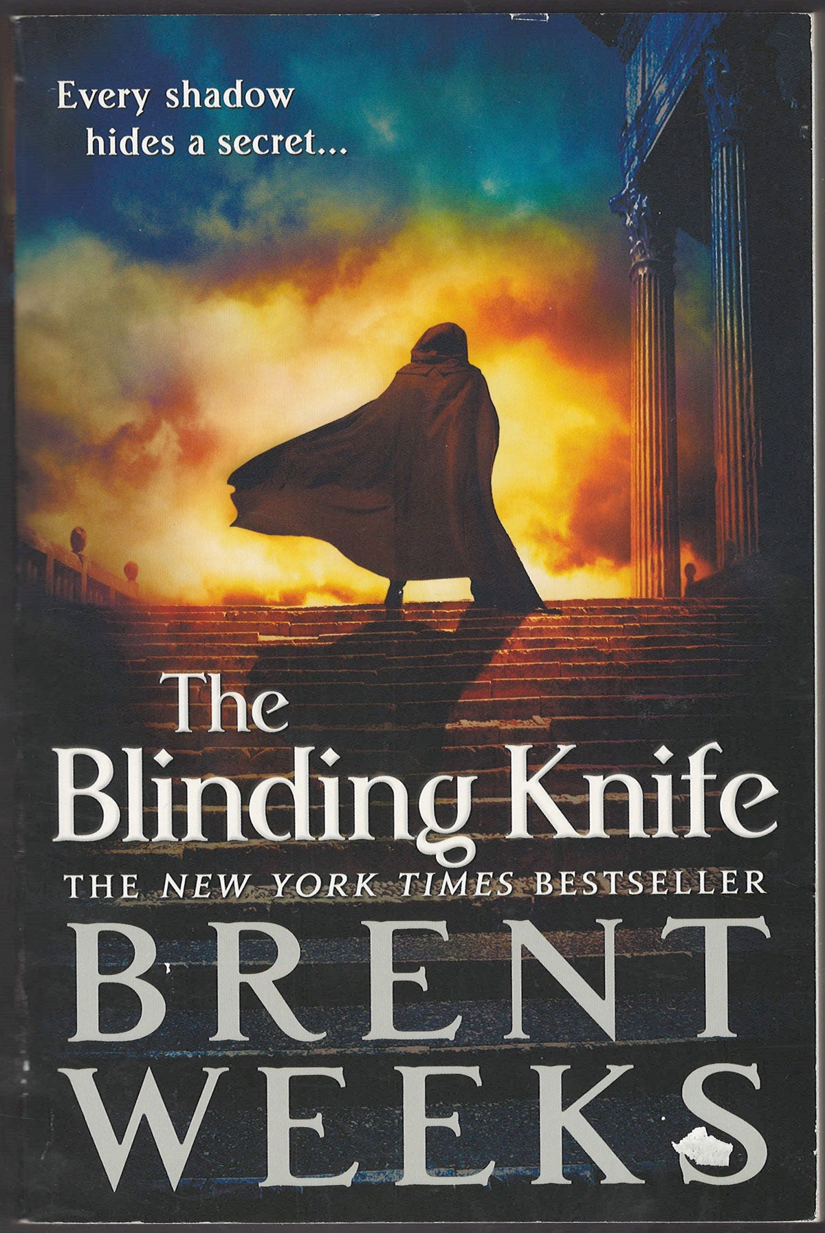 Front cover of The Blinding Knife by Brent Weeks with a silhouette of a cloaked figure walking up stairs into a fiery sky.