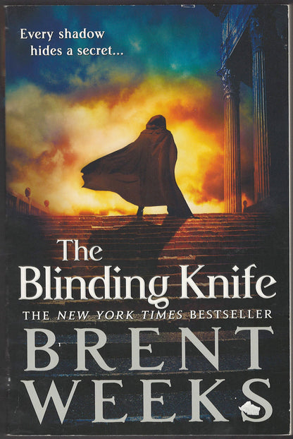 Front cover of The Blinding Knife by Brent Weeks with a silhouette of a cloaked figure walking up stairs into a fiery sky.
