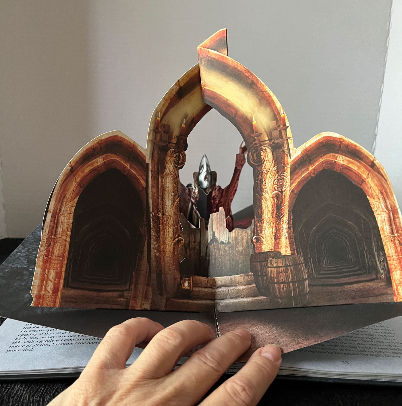 A hand holding open a 3D pop-up book showing a detailed scene of a ghostly figure standing between two large stone arches inside a tunnel.