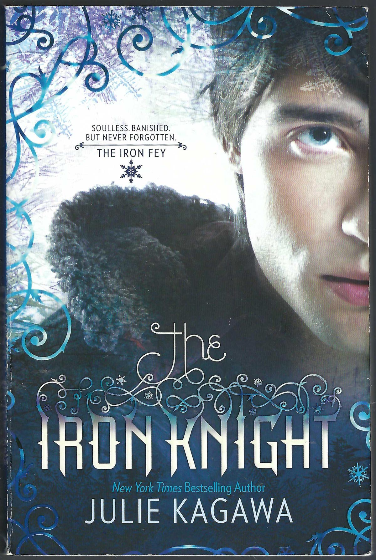 Front cover of The Iron Knight by Julie Kagawa, featuring a close-up portrait of a young man with blue eyes, a dark fur-lined hood, and icy blue decorative swirls on a light background.